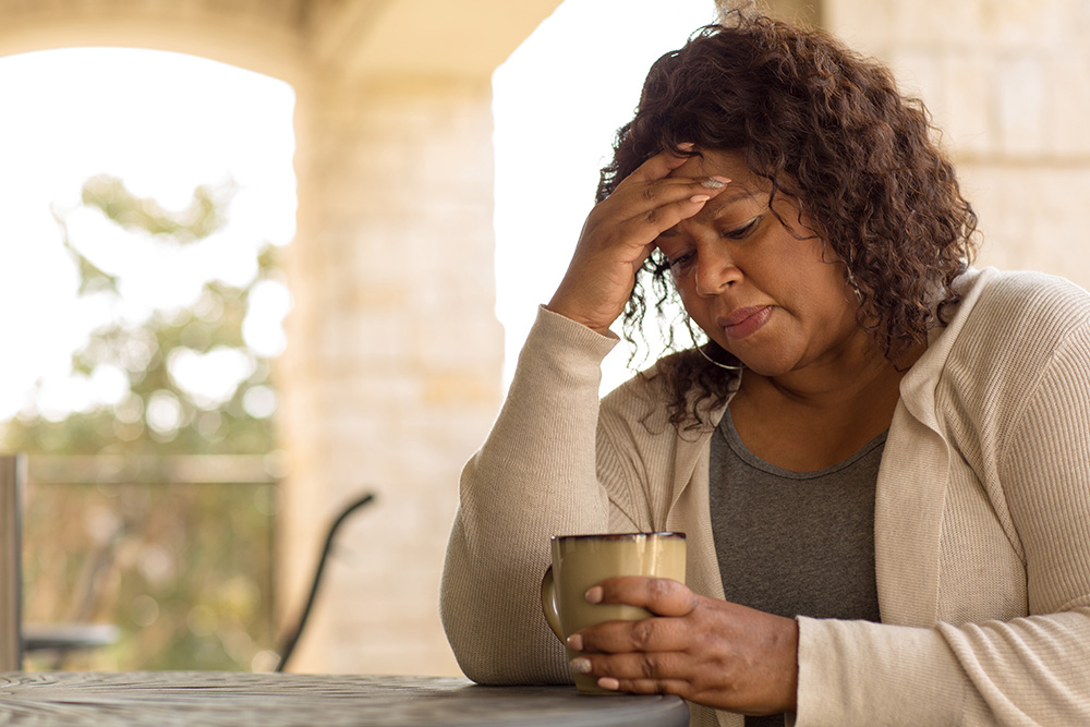 Middle-aged woman sits at kitchen table drinking tea and looking sad.